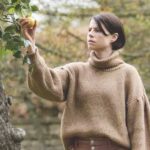 Filme Men - mulher colhendo uma maça amarela em uma árvore com expressão de curiosidade. Ao fundo arbustos desfocados.
