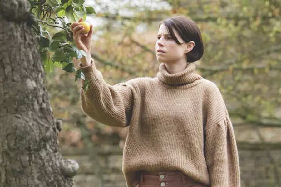 Filme Men - mulher colhendo uma maça amarela em uma árvore com expressão de curiosidade. Ao fundo arbustos desfocados.