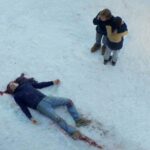 Anatomia de uma queda. Um homem caido sobre a neve manchada com seu sangue é observado pela esposa que segura seu filho com o rosto voltado para tras